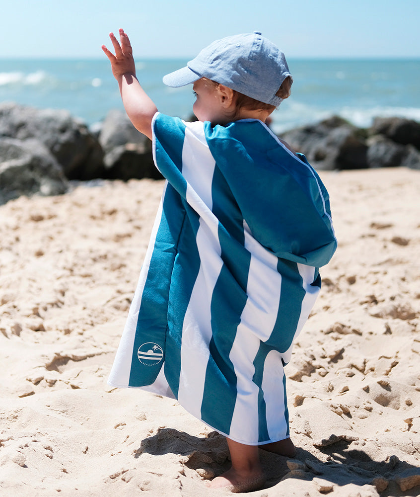 jeune enfant qui fait un signe de la main en regardant la mer et qui porte un poncho en microfibre