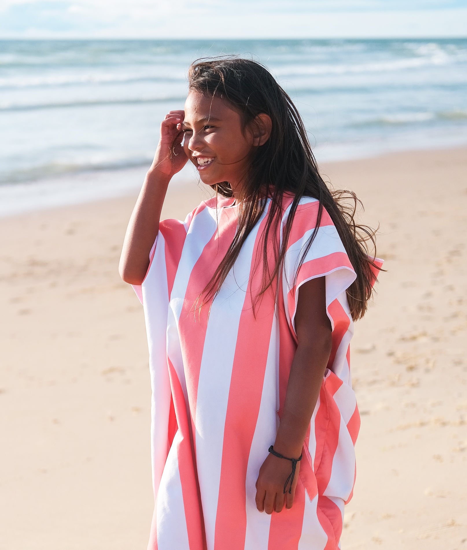 petite fille qui sourit sur la plage portant un poncho beachee couleur corail 