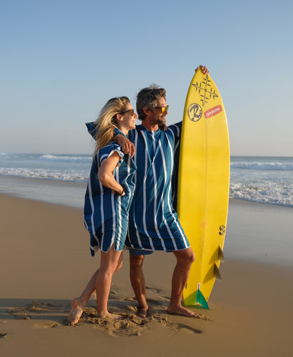 couple portant un poncho de plage et qui tiennent une planche de surf en regardant la mer