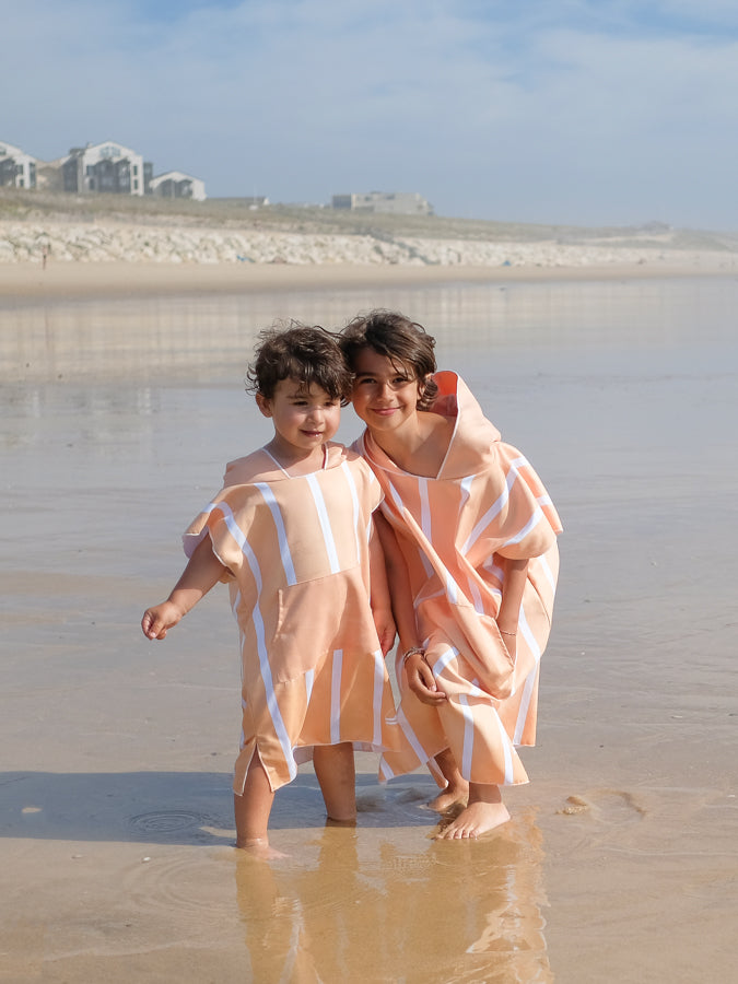 deux enfants avec les pieds dans l'eau en poncho couleur pêche 