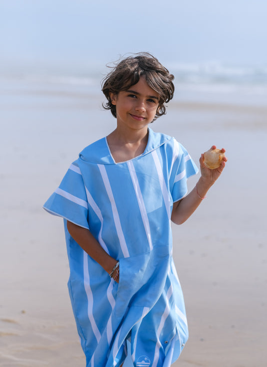 enfant qui tient un coquillage au bord de l'océan habillé en poncho bleu ciel